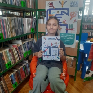 W bibliotece. Uśmiechnięta dziewczynka siedzi na pomarańczowej pufie. W ręku trzyma dyplom. W tle regały z książkami.