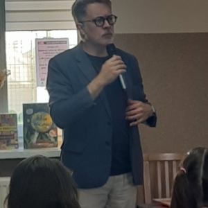 W bibliotece. Podczas spotkania autorskiego. Mężczyzna trzyma mikrofon, za nim na parapecie ustawione książki.