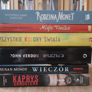 W bibliotece. Na stoliku ustawione książki. W tle regały z książkami.