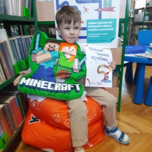 W bibliotece. Chłopiec siedzi na pomarańczowej pufie. W ręku trzyma dyplom i pluszową poduszkę. W tle regały z książkami.