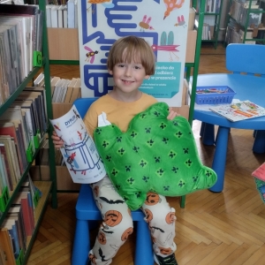 W bibliotece. Uśmiechnięty chłopiec siedzi na niebieskim krześle. W ręku trzyma dyplom i pluszową poduszkę w kolorze zielonym. W tle regały z książkami.