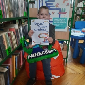 W bibliotece. Chłopiec siedzi na pomarańczowej pufie. W ręku trzyma dyplom. W tle regały z książkami.
