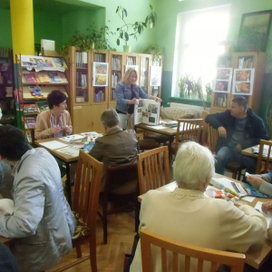 Uczestnicy spotkania siedzą przy stolikach i patrzą na panią prowadzącą, która pokazuje największą książkę dostępną w bibliotece.