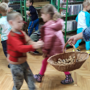 Dzieci zbierają orzechy do koszyka, który w ręku trzyma kobieta.