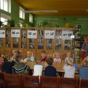 Wnętrze biblioteki. W tle regały z książkami. Przy długim stole siedzi grupa dzieci.