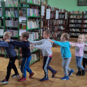 Wnętrze biblioteki. W tle regały z książkami. Dzieci bawią się w tzw. węża.