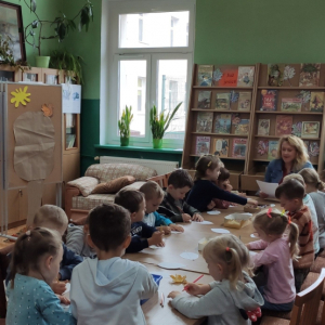 Wnętrze biblioteki.  Przy długim stole siedzi grupa dzieci. Dzieci kolorują liście i słuchają pani, która im czyta wiersze i zagadki.