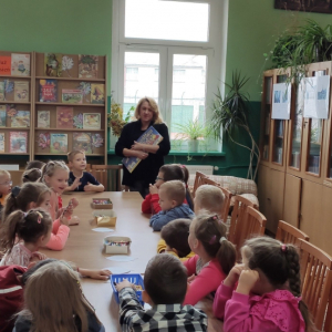 Wnętrze biblioteki. W tle regały z książkami. Przy długim stole siedzi grupa dzieci, która słucha pani prowadzącej spotkanie.