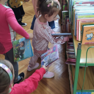 Wnętrze biblioteki. W tle regały z książkami. 5 dzieci stoi przy zielonych regałach i przegląda książki.