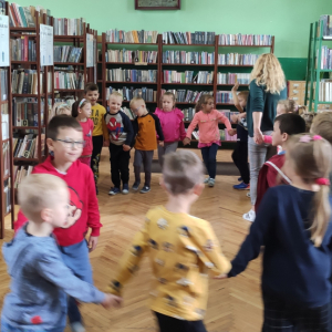 Wnętrze biblioteki. W tle regały z książkami. Dzieci ustawiły się w kółeczku i wspólnie tańczą.. 