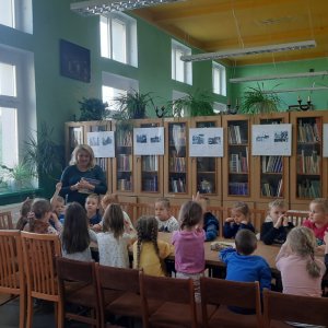 Wnętrze biblioteki. W tle regały z książkami. Przy długim stole siedzi grupa dzieci