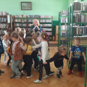 Wnętrze biblioteki. W tle regały z książkami. Dzieci ustawiły się w kółeczku i wspólnie tańczą.