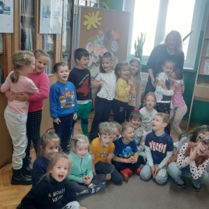 Wnętrze biblioteki. Dzieciu stoją przy korkowym parawanie, na którym wywieszona jest praca plastyczna, którą wspólnie wykonali. W tle gablota wystawowa i przeszklone regały z książkami.