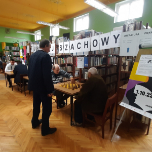 W pomieszczeniu biblioteki. Mężczyźni siedzą przy stolikach i grają w szachy. Przy stoliku stoi pan z Komisji i obserwuje rozgrywkę. W tle regały z książkami.