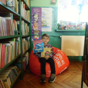 W pomieszczeniu biblioteki. Chłopiec siedzi na pomarańczowej pufie. W ręku trzyma dyplom i książkę. W tle regały z książkami