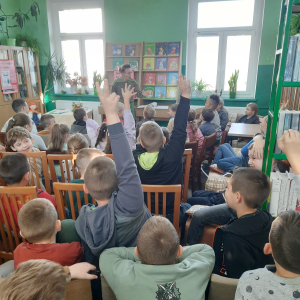 W pomieszczeniu biblioteki. Pani prowadząca spotkanie rozmawia z dziećmi. Dzieci, które chcą zadać pytanie trzymają rękę w górze.