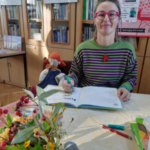 W pomieszczeniu biblioteki. Kobieta siedzi przy stoliku, uśmiecha się, w ręku trzyma mazak i podpisuje książkę.