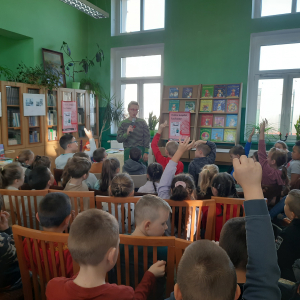 W pomieszczeniu biblioteki. Pani prowadząca spotkanie trzyma  mikrofon w ręku. Dzieci, które chcą zadać pytanie mają podniesioną rękę do góry.