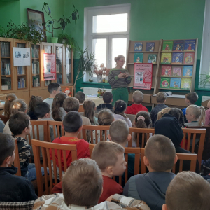 W pomieszczeniu biblioteki. Grupa dzieci słucha kobiety, która prowadzi spotkanie.