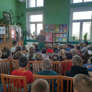 W pomieszczeniu biblioteki. Grupa dzieci słucha kobiety, która prowadzi spotkanie. Kobieta trzyma w ręku książkę.