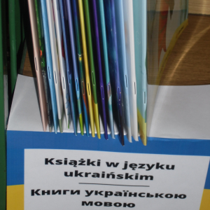 Na półce ustawione pionowo książki, grzbietem do przodu. Niżej flaga Ukrainy, na niej napis w języku polskim i ukraińskim.