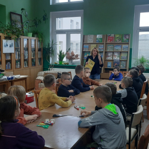 W bibliotece. Grupa dzieci siedzi przy stolikach. W tle regały z książkami.