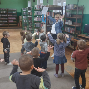 W pomieszczeniu biblioteki. Grupa dzieci uczestniczy w zajęciach ruchowych.