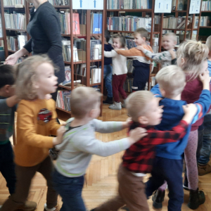 "W pomieszczeniu biblioteki. Grupa dzieci uczestniczy w zajęciach ruchowych.