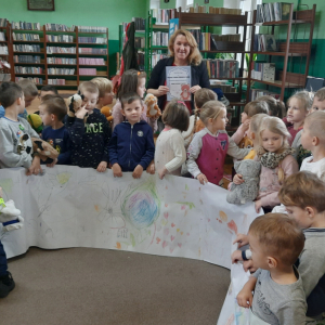 W bibliotece. W tle regały z książkami. Dzieci trzymają dużą laurkę, którą wspólnie wykonały.