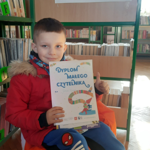 W pomieszczeniu biblioteki. Uśmiechnięty chłopiec siedzi na pomarańczowej pufie. W ręku trzyma dyplom. W tle regały z książkami.