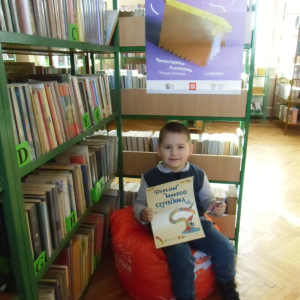 W pomieszczeniu biblioteki. Uśmiechnięty chłopiec siedzi na pomarańczowej pufie. W ręku trzyma dyplom. W tle regały z książkami.