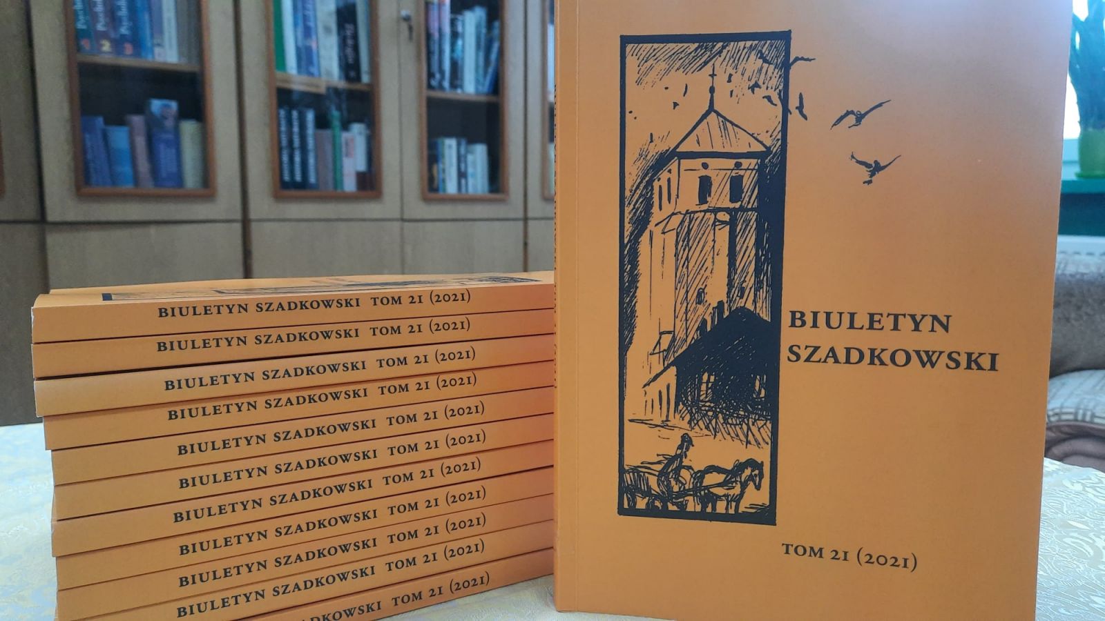 11 egzemplarzy "Biuletynu Szadkowskiego" ułożone na stoliku jeden na drugim, grzbietem okładki do przodu. Jedna książka ustawiona pionowo stroną tytułową. W tle widać oszklone regały z książkami
