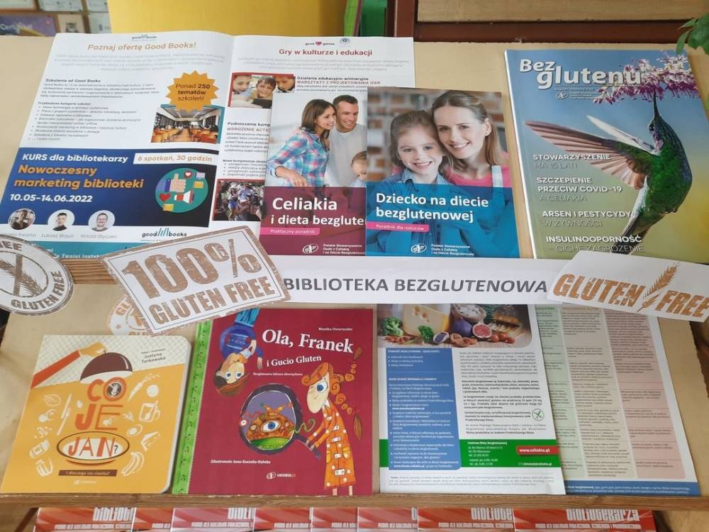 "Wystawa książek i broszur otrzymanych w ramach nagrody za udział w konkursie \"Biblioteka bezglutenowa\". Książki i broszurki ustawione jedna obok drugiej, przodem okładki. Na środku wydrukowany napis Biblioteka bezglutenowa i 100% gluten free"